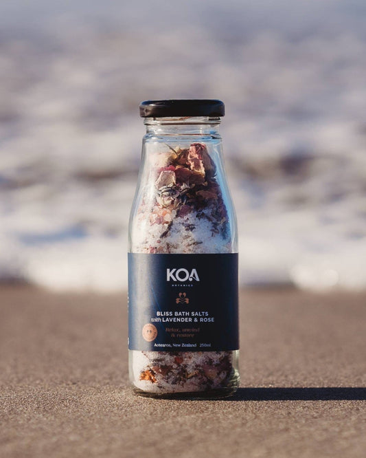 Koa Bliss Bath Salts with Lavender & Rose sitting on the sand with the ocean behind.