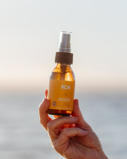 A hand holding a small amber spray bottle labeled KOA Natural Hair Reviver Oil against a soft, blurred beach background at sunset.