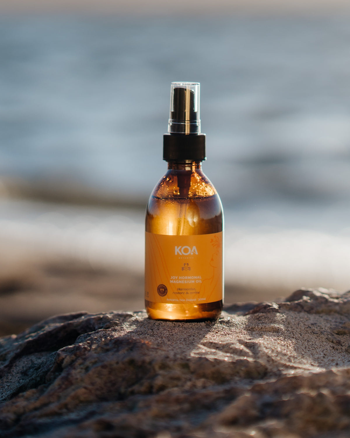 An amber glass spray bottle with a black pump and clear cap stands on a rock outdoors, with a blurred beach or ocean background. The label shows the brand KOA.