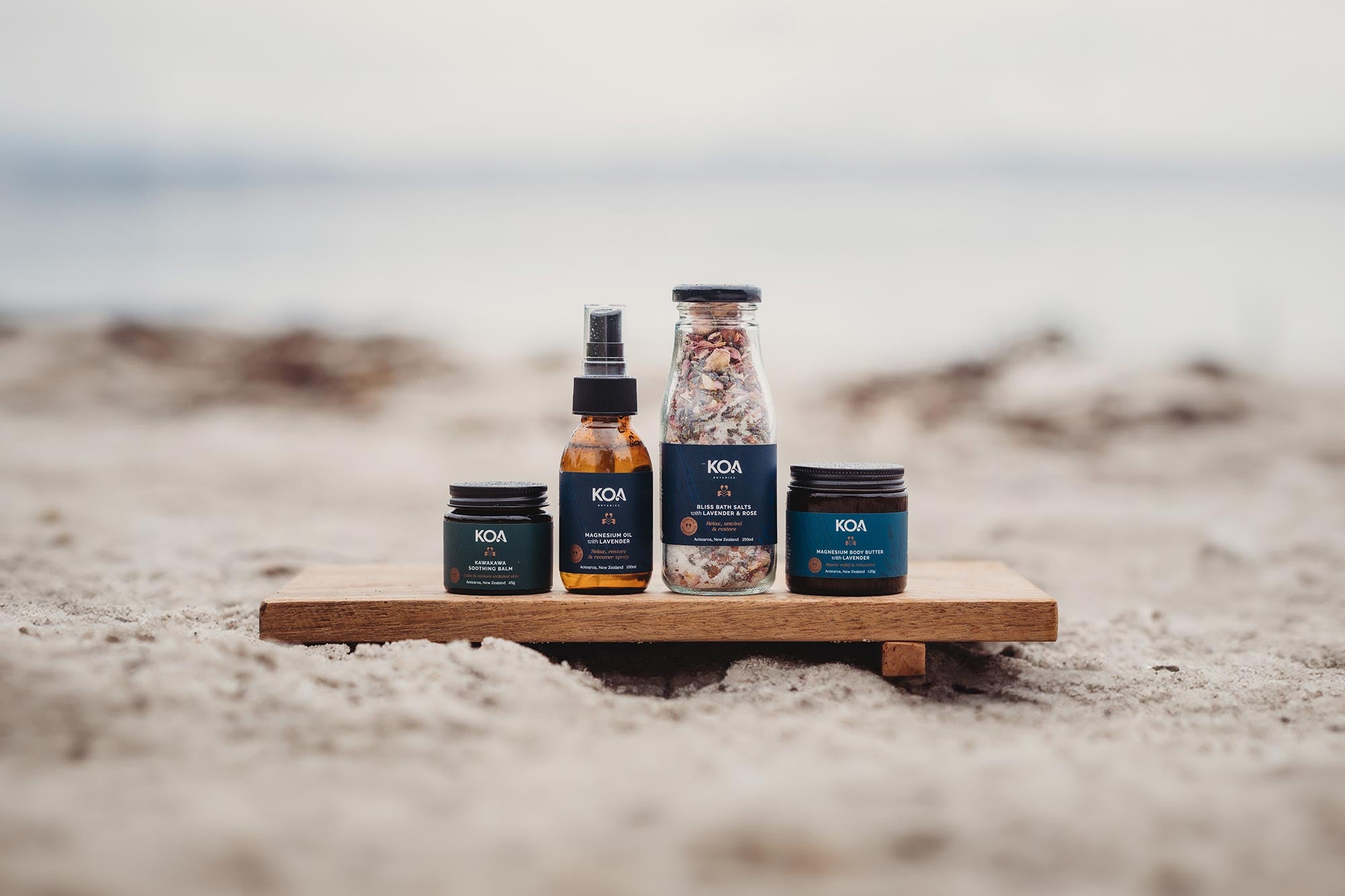 Four Koa Botanics products sitting on a wooden plinth on the sand with a beach background.