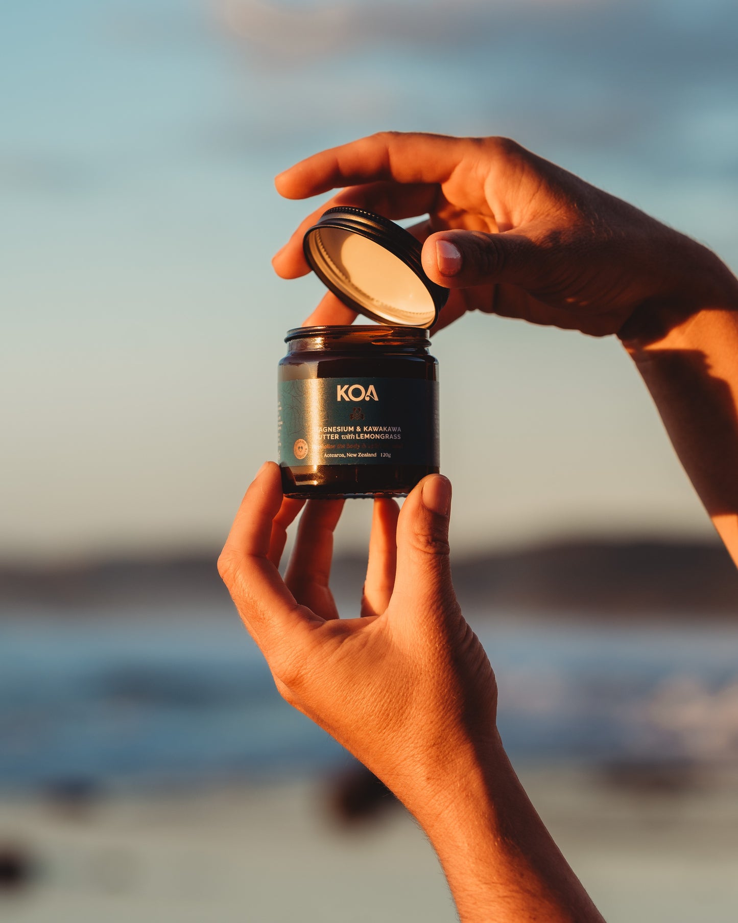 Hands holding Magnesium Butter with Kawakawa and Lemongrass, opening lid against an ocean background.
