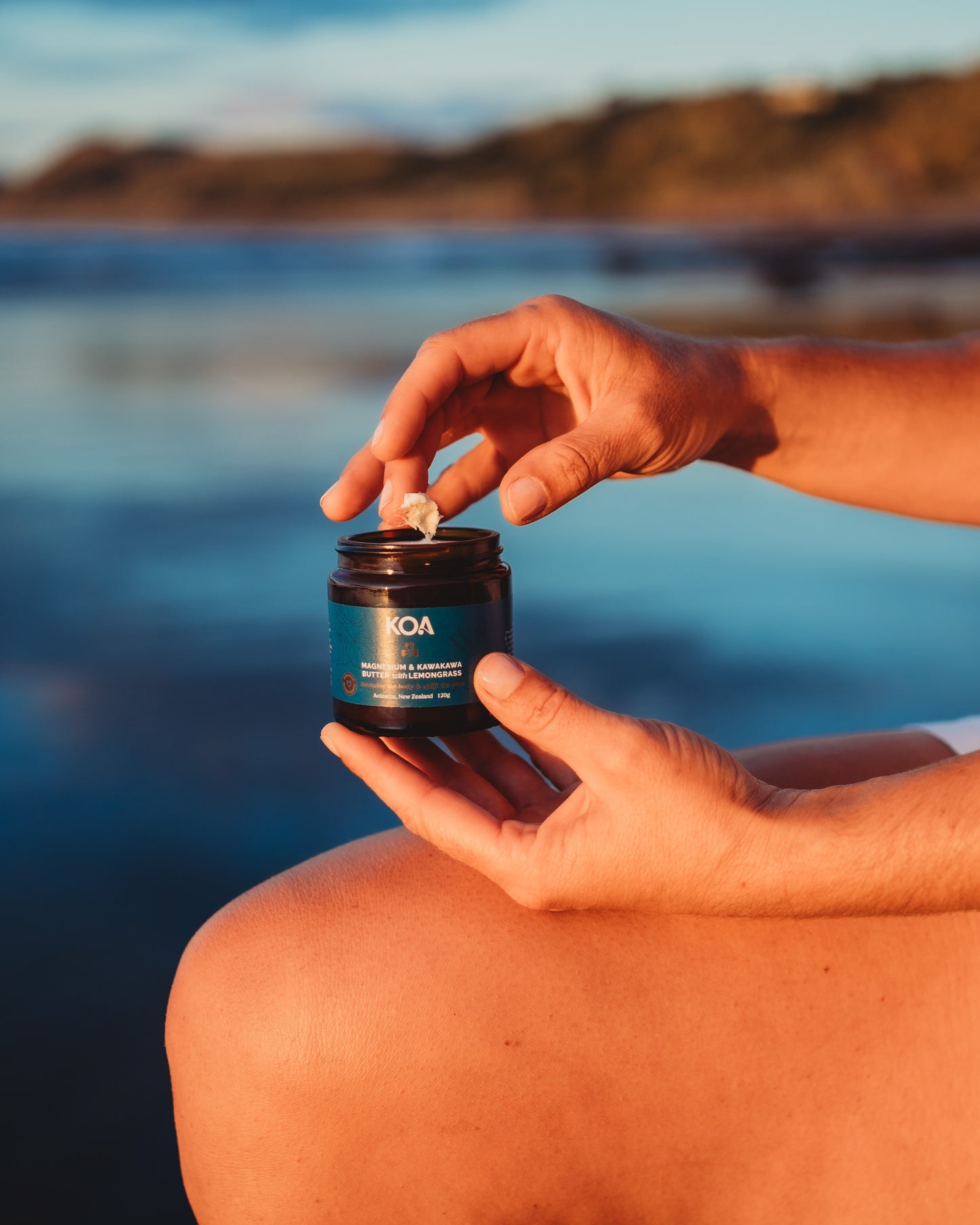 An open jar of Magnesium Butter with hands sampling the content with beach background.