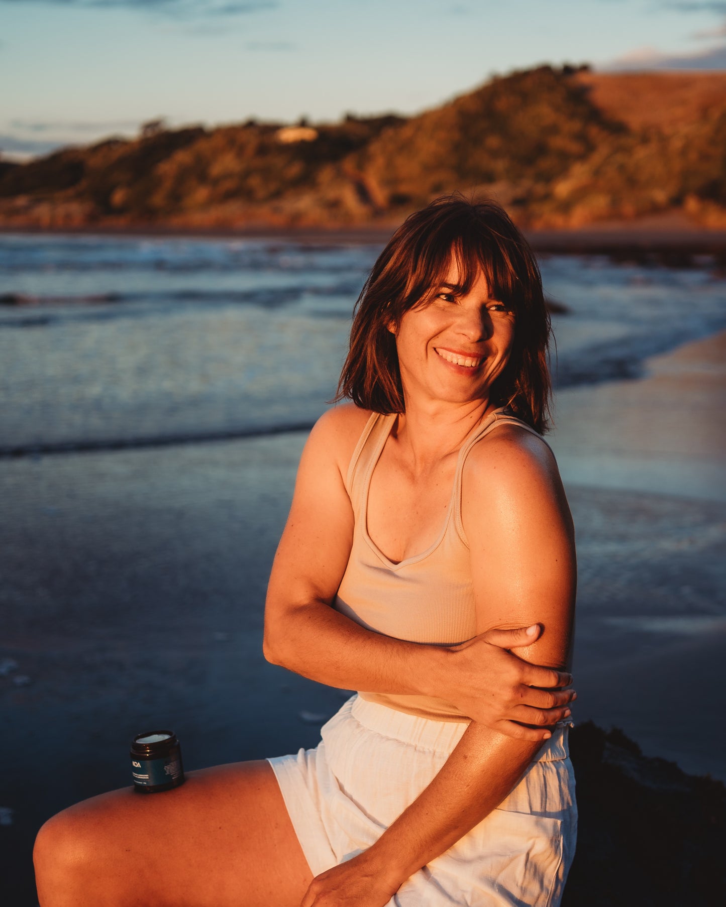 Smiling woman sitting on a rock at the beach applying Magnesium Butter to her arm.