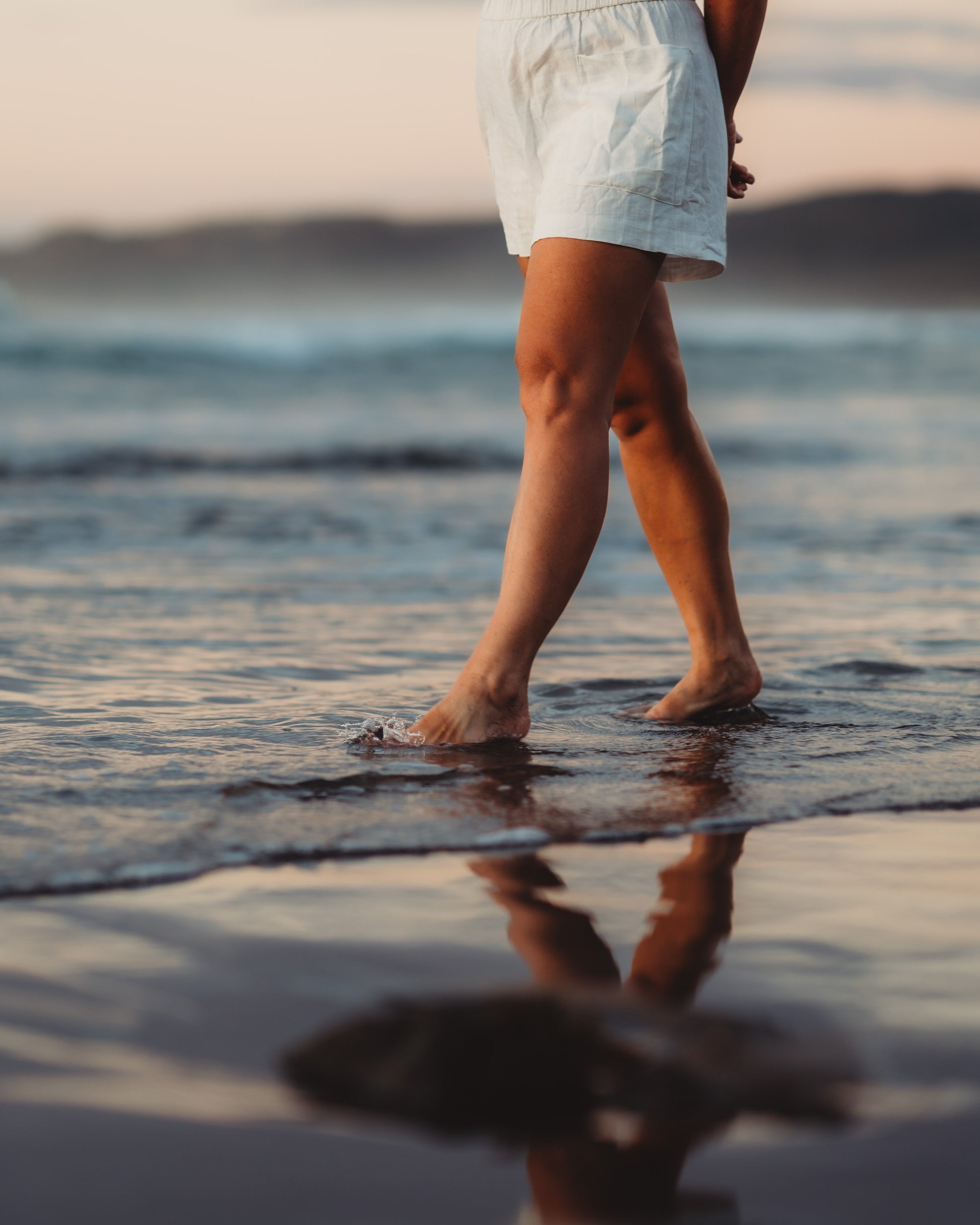 Lower half of a woman walking through the water at the shoreline with an ocean background.