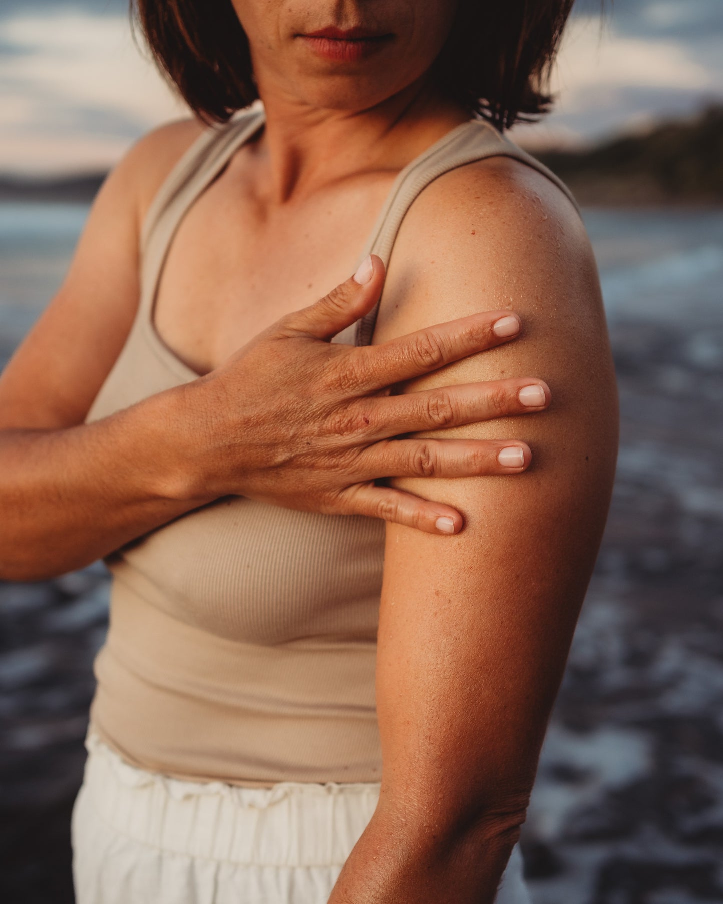 Female showing lower face and upper body with her hand on upper arm and an ocean background.