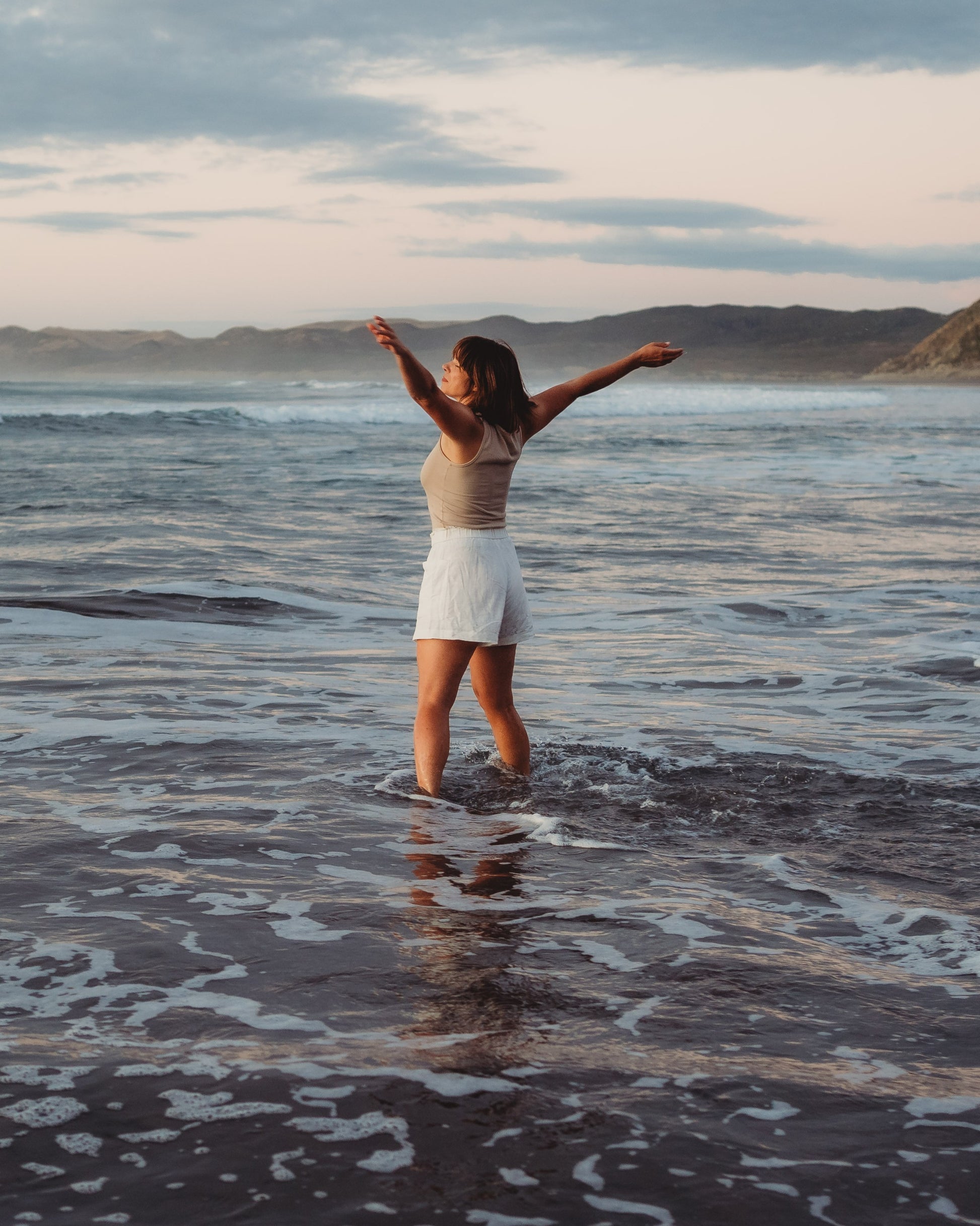 Woman standing in the ocean with arms in the air looking out to the ocean.