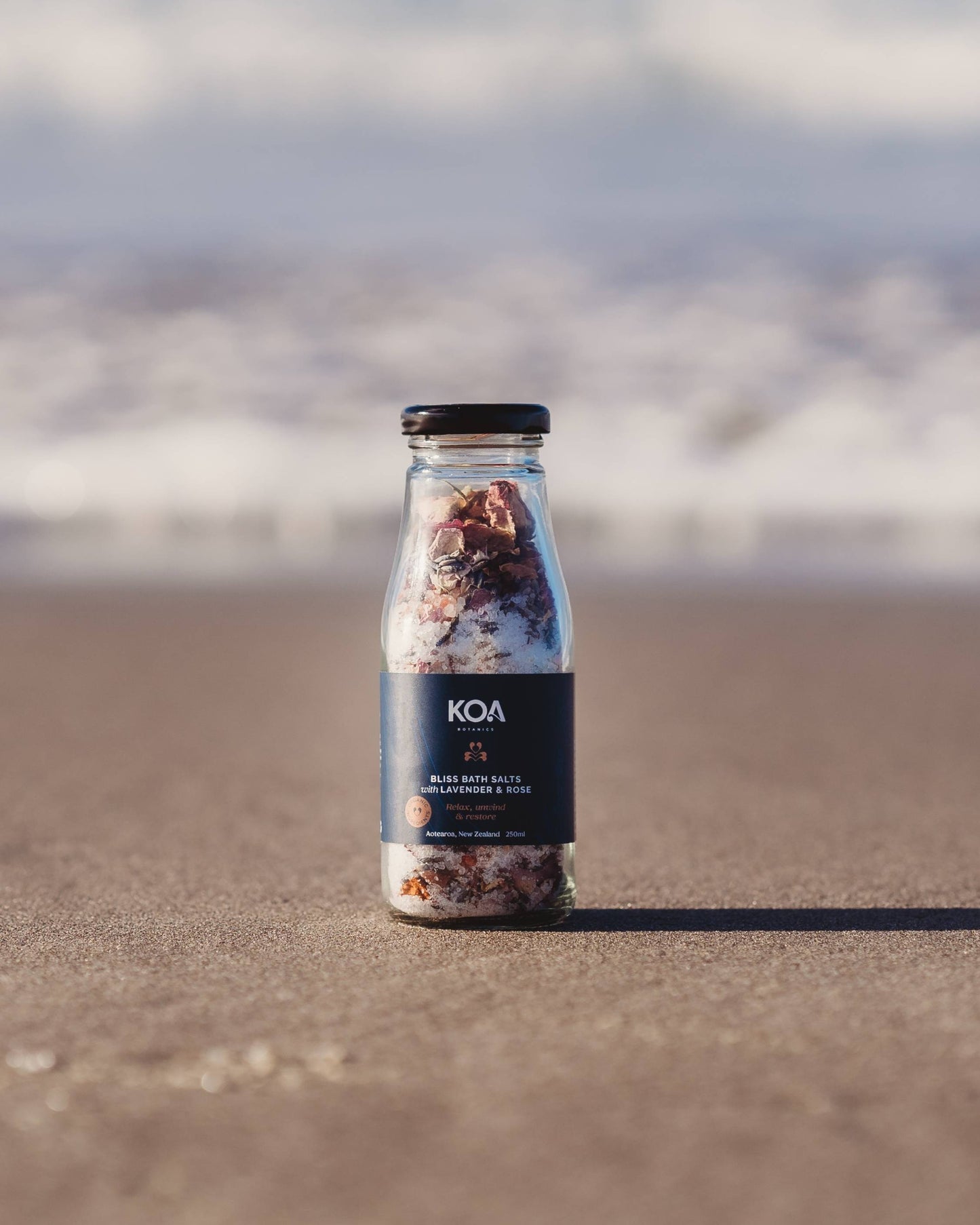 Koa Bliss Bath Salts with Lavender & Rose sitting on the sand with the ocean behind.