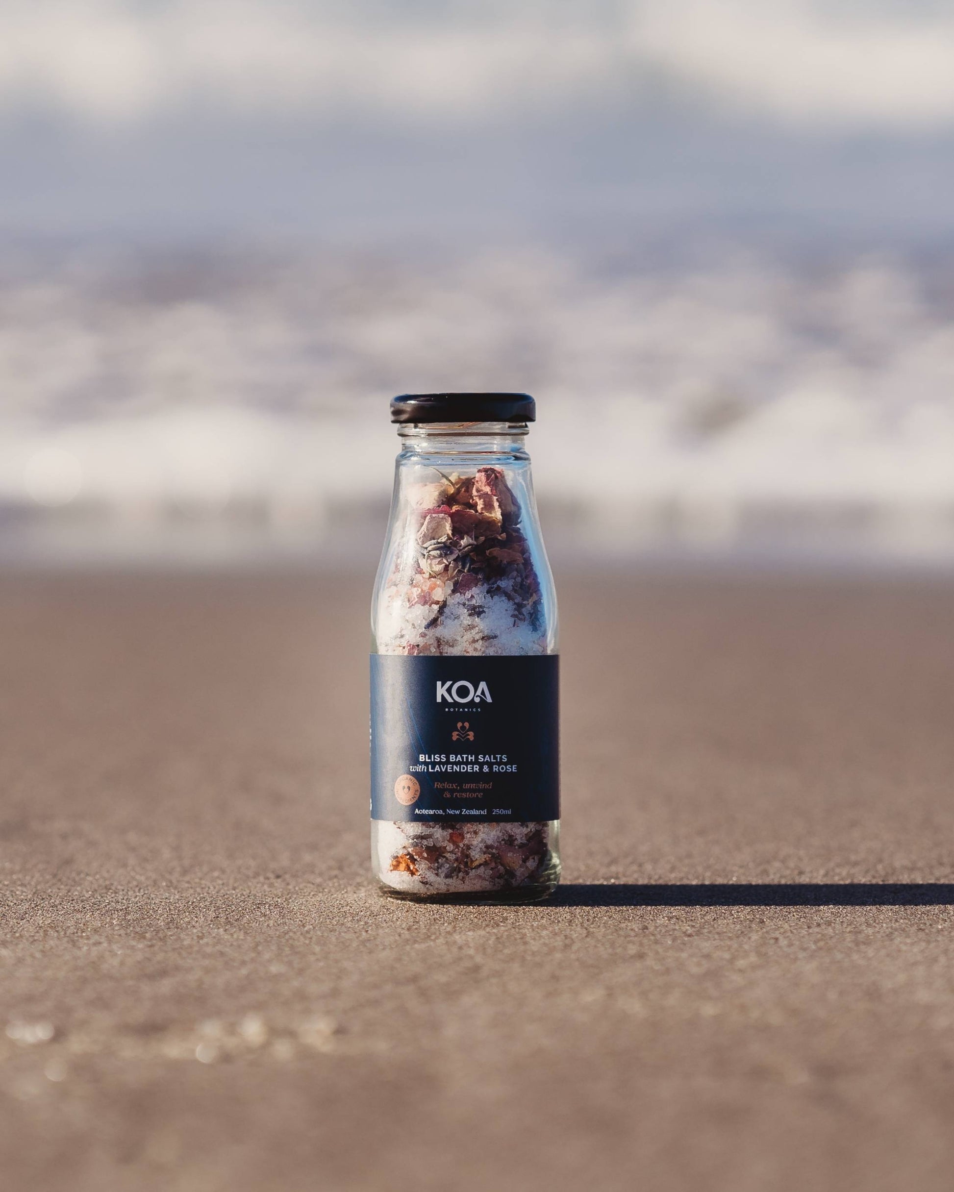 Koa Bliss Bath Salts with Lavender & Rose sitting on the sand with the ocean behind.