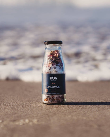 Koa Bliss Bath Salts with Lavender & Rose sitting on the sand with the ocean behind.