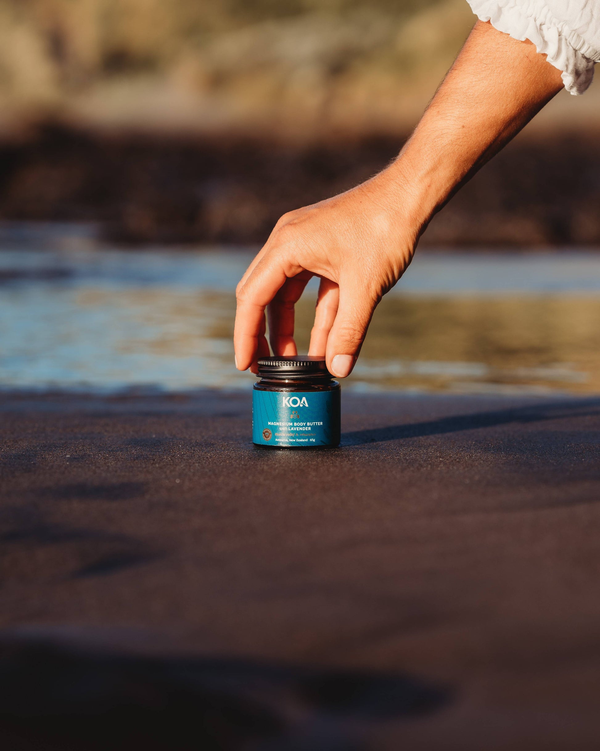 Magnesium Butter with Lavender sitting on the sand with hand reaching down touching the lid from above.