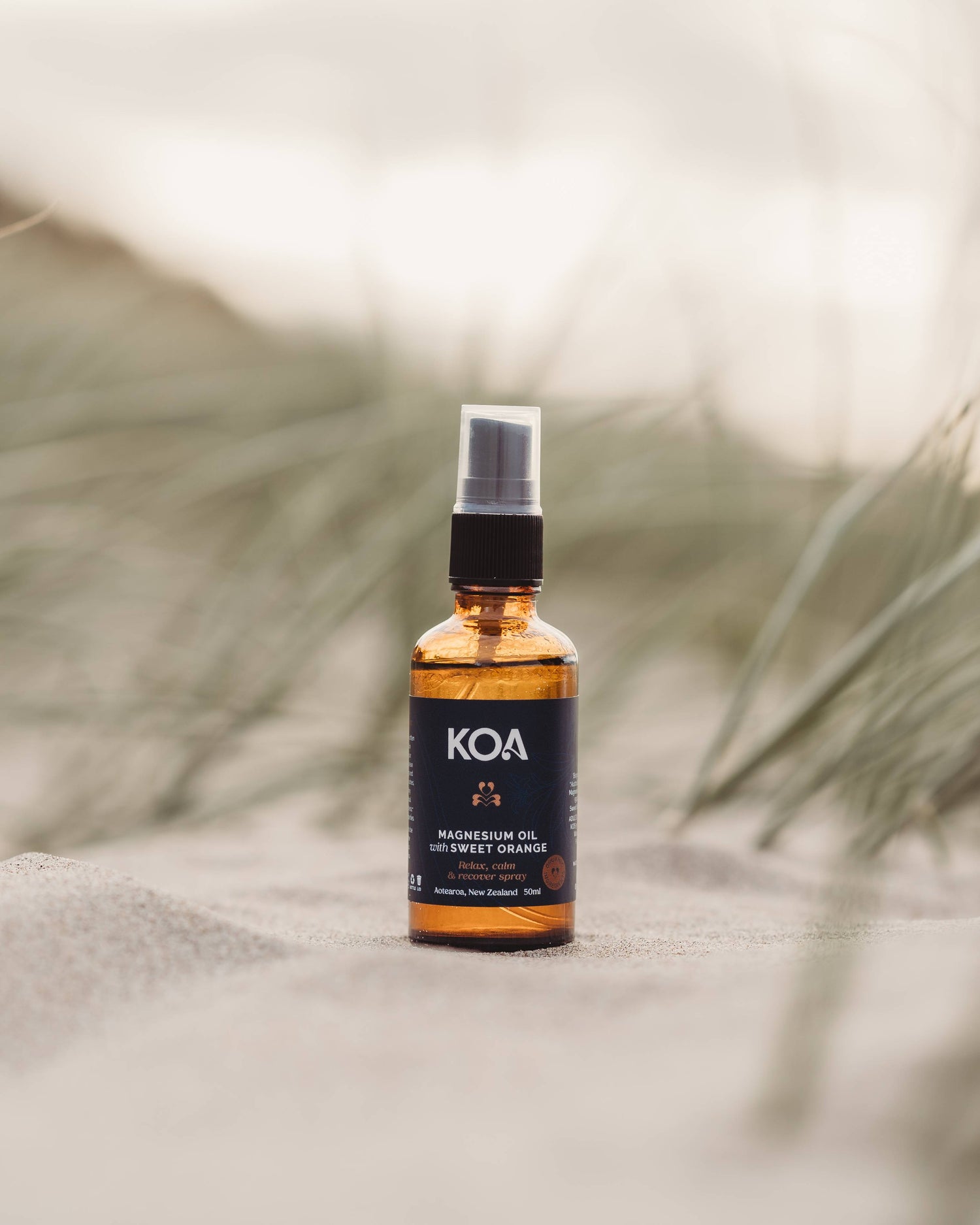 Magnesium Oil with Sweet Orange sitting on the sand with grasses behind.
